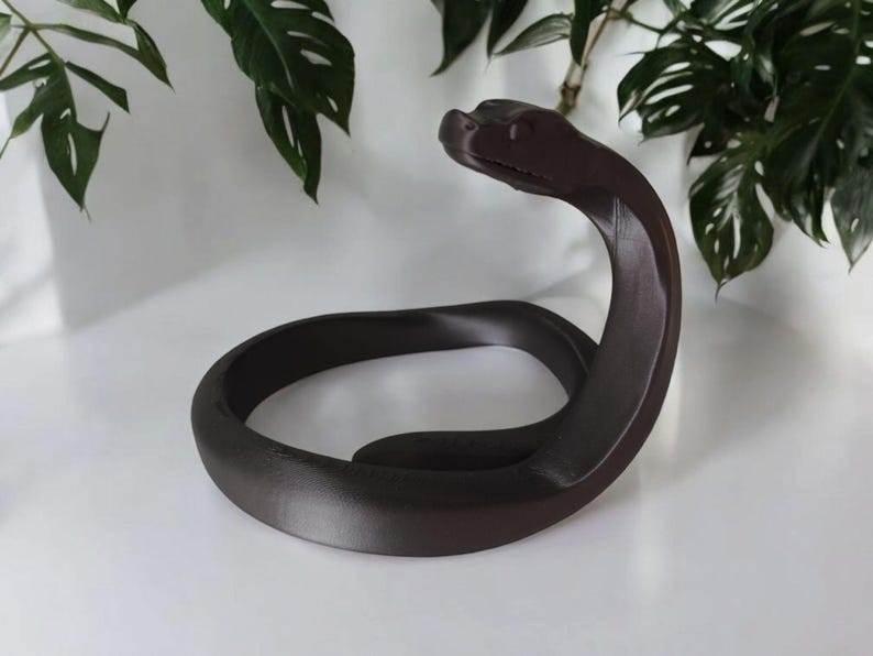 Puede incluir: Una escultura de serpiente marr&oacute;n oscuro con un cuerpo curvado y la cabeza levantada. La escultura est&aacute; hecha de un material liso y se encuentra sobre una superficie blanca. Se ven hojas verdes en el fondo.