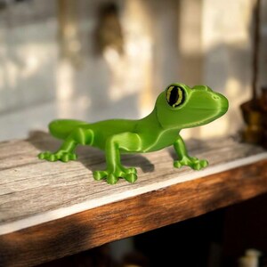 May include: A bright green, 3D-printed gecko figurine. The lizard has a detailed design with visible legs, a long tail, and large eyes with black pupils. The gecko is posed on a wooden surface, showcasing its vibrant color and intricate features.