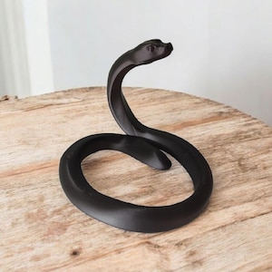 Puede incluir: Una escultura de serpiente negra y estilizada, con un cuerpo curvado y la cabeza levantada. La escultura est&aacute; hecha de un material mate y se encuentra sobre una mesa redonda de madera. La forma de la serpiente es suave y elegante.
