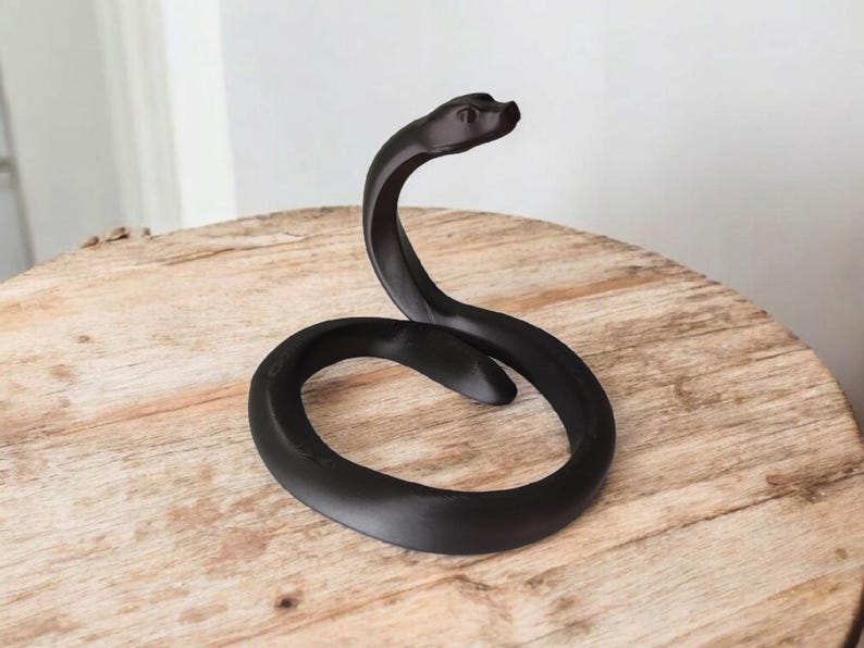 Puede incluir: Una escultura de serpiente negra y estilizada, con un cuerpo curvado y la cabeza levantada. La escultura est&aacute; hecha de un material mate y se encuentra sobre una mesa redonda de madera. La forma de la serpiente es suave y elegante.