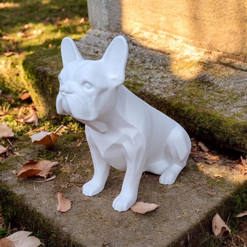 May include: White ceramic sculpture of a French bulldog sitting on a stone surface. The sculpture is detailed and realistic, with a smooth, matte finish.