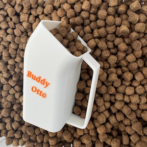 Może przedstawiać: Biała plastikowa łopatka z nadrukiem "Buddy Otto" w kolorze pomarańczowym. Łopatka jest wypełniona brązowymi karmą dla ps&oacute;w.