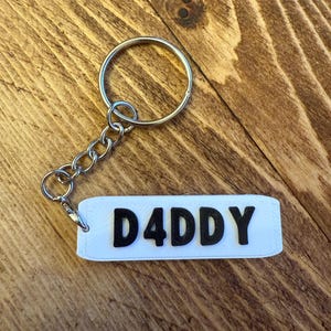 May include: A white rectangular keychain with the text "D4DDY" in black block letters. The keychain is attached to a silver metal split ring and chain. The background is a wooden surface.