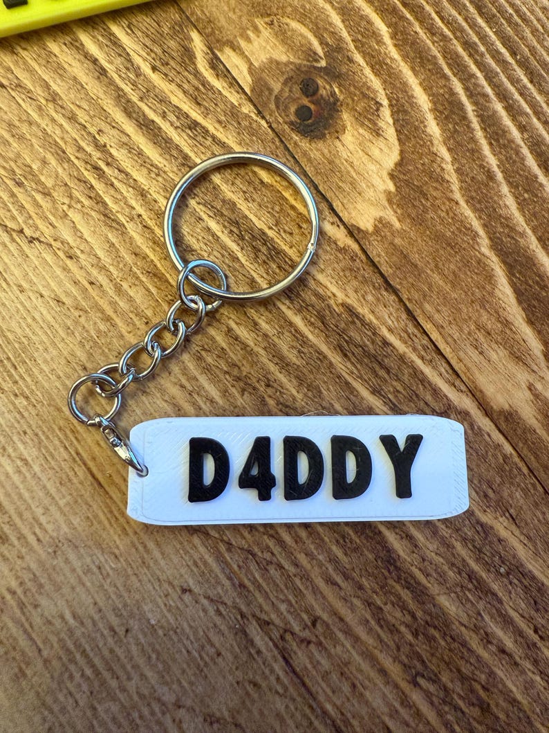 May include: A white rectangular keychain with the text "D4DDY" in black block letters. The keychain is attached to a silver metal split ring and chain. The background is a wooden surface.