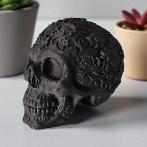 May include: A black decorative skull with intricate floral patterns. The skull is a detailed sculpture, showcasing a realistic depiction of the human skull with a decorative overlay. The skull is a dark matte black color.