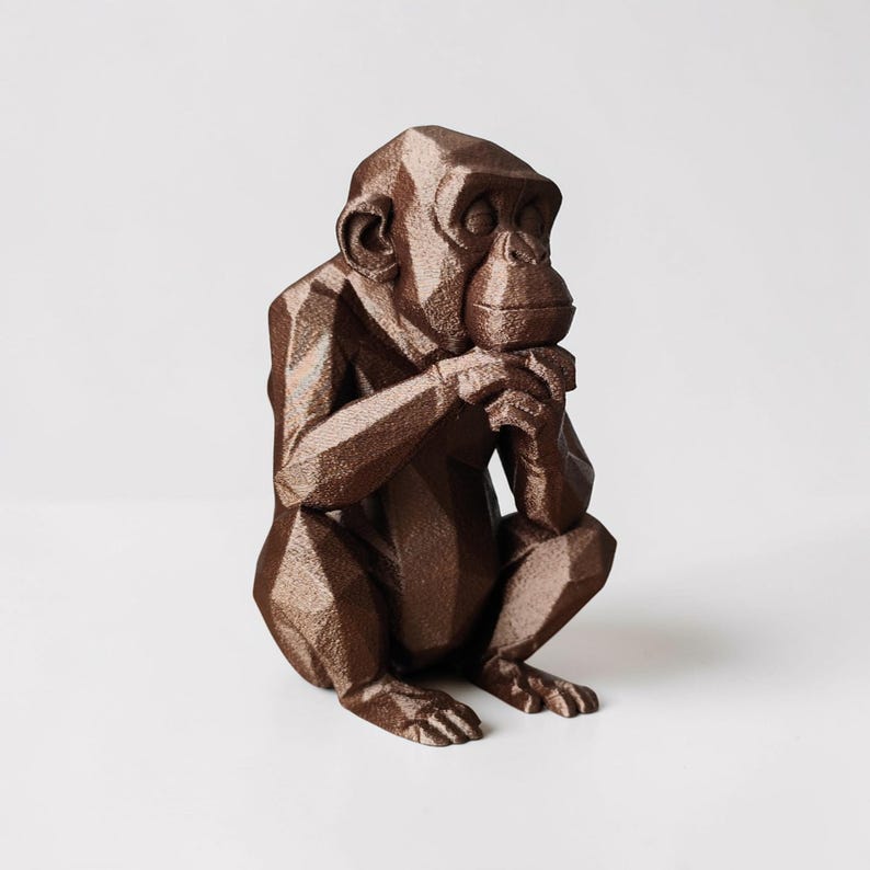 May include: A brown geometric chimpanzee sculpture in a seated position, with hands clasped near its mouth. The sculpture has a faceted design, giving it a modern, angular appearance. The chimpanzee is on a white background.