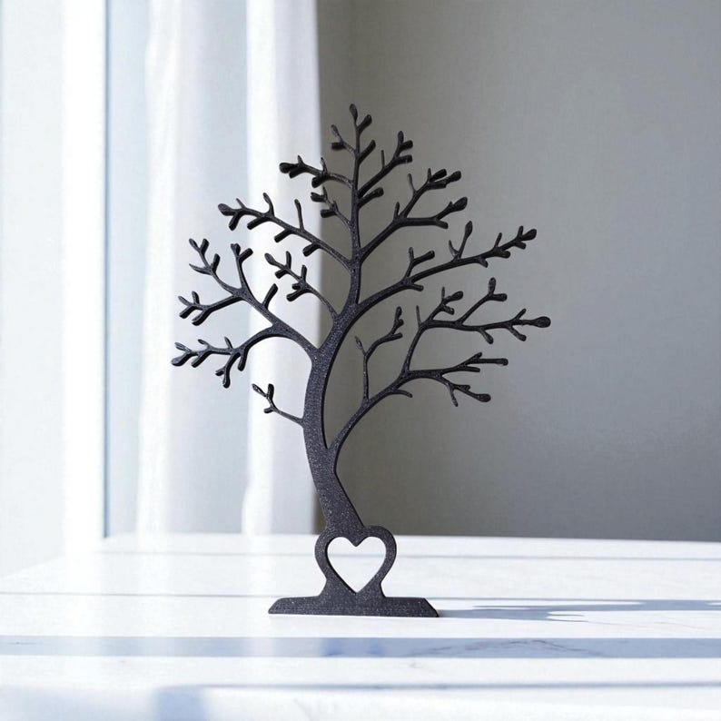 May include: A black metal jewelry tree in the shape of a tree with a heart-shaped cutout. The tree has many branches for hanging jewelry and sits on a white surface. The background is a white curtain and a gray wall.