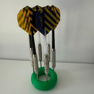 May include: A collection of darts with yellow and navy blue striped flights, black shafts, and silver metal tips. The darts are held in a green circular holder. The flights are printed with the word "NUKE".