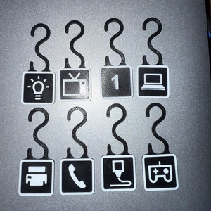 May include: Eight black and white 3D-printed hooks with various icons. The icons include a lightbulb, television, the number one, a laptop, a printer, a phone, a charging cable, and a game controller.