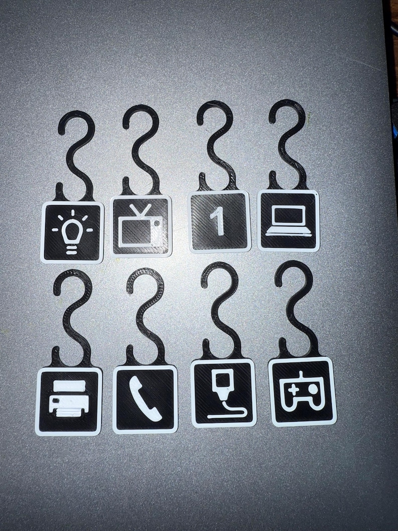 May include: Eight black and white 3D-printed hooks with various icons. The icons include a lightbulb, television, the number one, a laptop, a printer, a phone, a charging cable, and a game controller.
