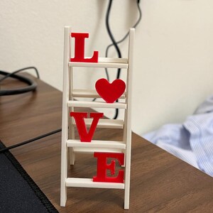 May include: A small, white wooden ladder with the word "LOVE" spelled out in red letters and a red heart. The decorative piece is on a brown wooden surface.