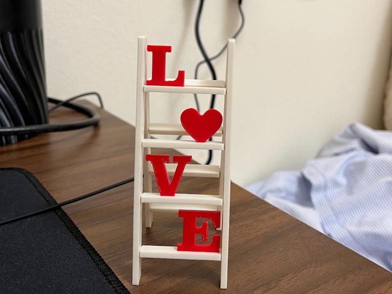 May include: A small, white wooden ladder with the word "LOVE" spelled out in red letters and a red heart. The decorative piece is on a brown wooden surface.