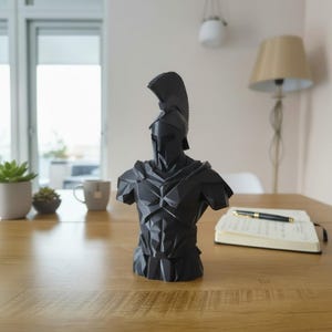 May include: A black Spartan warrior bust, with a helmet and detailed armour. The sculpture is on a wooden table, with a notebook and pen. The bust is approximately 30 cm tall.