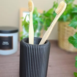 May include: A black, ribbed toothbrush holder with two bamboo toothbrushes. The toothbrushes have light-colored bristles and are placed inside the holder. A candle with the text "FRESH COTTON" is in the background.