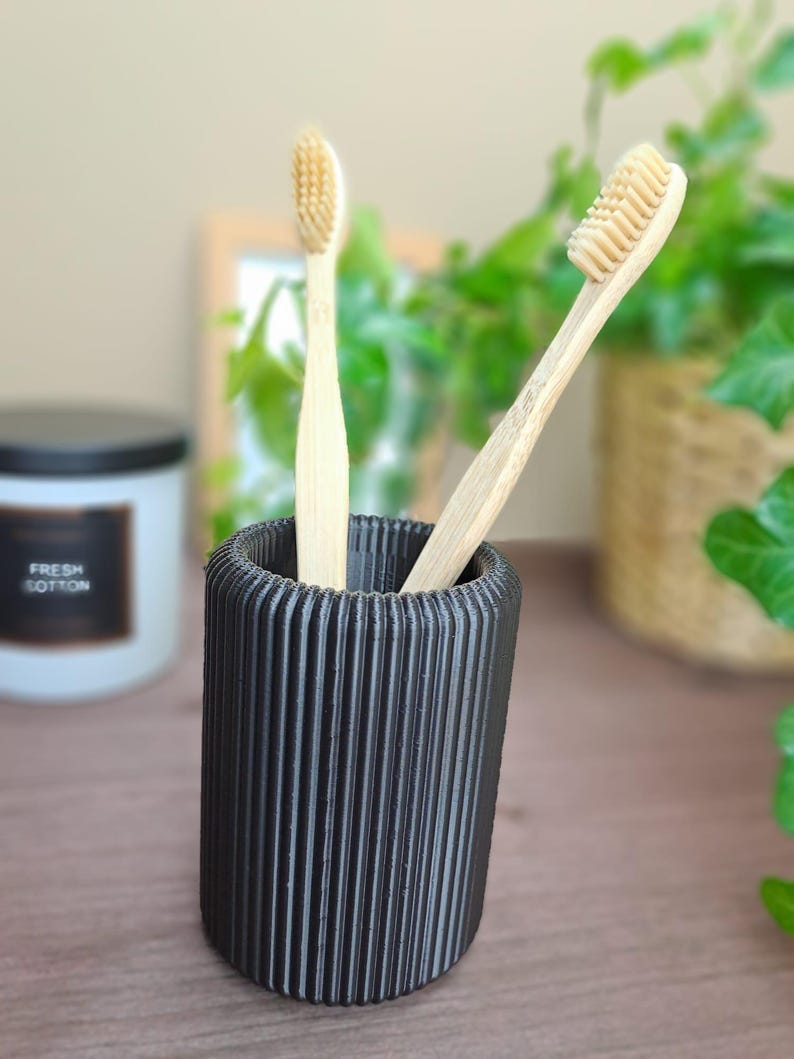 May include: A black, ribbed toothbrush holder with two bamboo toothbrushes. The toothbrushes have light-colored bristles and are placed inside the holder. A candle with the text "FRESH COTTON" is in the background.