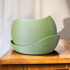 May include: A textured, sage green ceramic plant pot with a matching saucer. The rounded pot has a unique, curved design. The pot and saucer sit on a wooden surface, creating a natural aesthetic. Ideal for indoor plants.