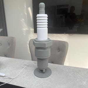 May include: A large, gray and white decorative spark plug sculpture. The sculpture is made of several cylindrical and hexagonal components, with a rounded top and a circular base. The spark plug is displayed on a gray surface.