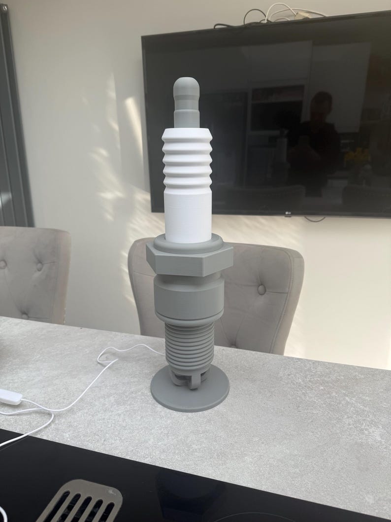 May include: A large, gray and white decorative spark plug sculpture. The sculpture is made of several cylindrical and hexagonal components, with a rounded top and a circular base. The spark plug is displayed on a gray surface.