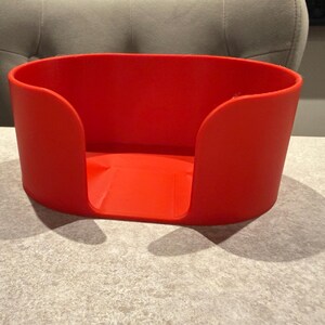 May include: A bright red, oval-shaped container with curved sides and a flat base. The container is made of a solid material and sits on a light gray surface. The design features a cutout on one side.