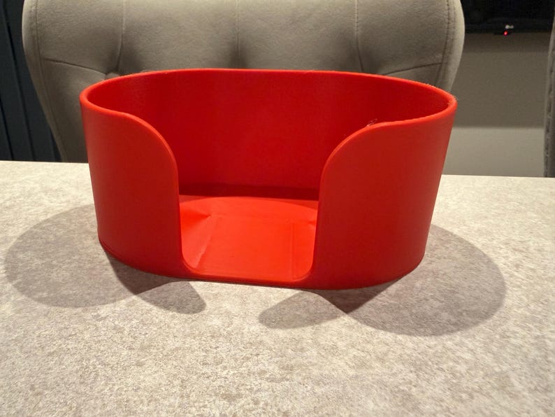 May include: A bright red, oval-shaped container with curved sides and a flat base. The container is made of a solid material and sits on a light gray surface. The design features a cutout on one side.
