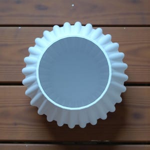 May include: Overhead view of a white, ruffled, circular object with a gray interior. The object is set against a wooden background. The ruffled edge has a repeating pattern, and the interior is smooth.