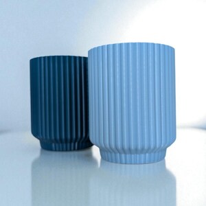 May include: Two cylindrical vases with vertical ribbed detailing. One vase is a deep teal color, and the other is a light blue. The vases have a rounded base and a slightly flared top. They are sitting on a white surface.