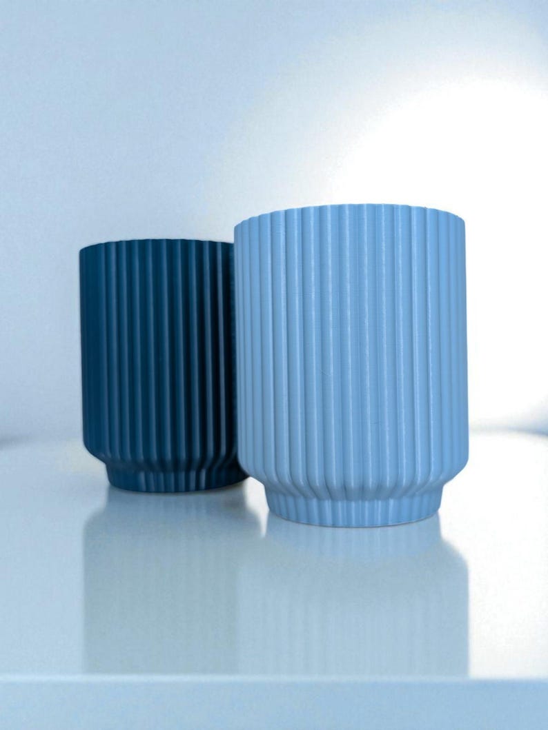 May include: Two cylindrical vases with vertical ribbed detailing. One vase is a deep teal color, and the other is a light blue. The vases have a rounded base and a slightly flared top. They are sitting on a white surface.