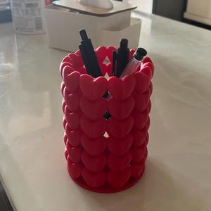 Puede incluir: Un portal&aacute;pices rojo con forma de corazones apilados, que contiene varios bol&iacute;grafos negros y un l&aacute;piz. El portal&aacute;pices es cil&iacute;ndrico y est&aacute; sobre una superficie blanca. Una caja de pa&ntilde;uelos blanca est&aacute; en el fondo.