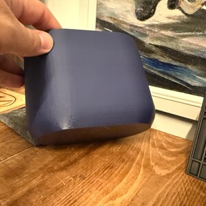 May include: A solid, navy blue, square-shaped container with rounded edges. The container is held by a hand and sits on a wooden surface. A wooden coaster and a black plastic object are also visible in the background.