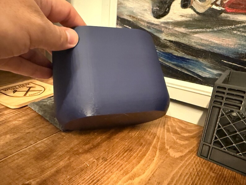 May include: A solid, navy blue, square-shaped container with rounded edges. The container is held by a hand and sits on a wooden surface. A wooden coaster and a black plastic object are also visible in the background.
