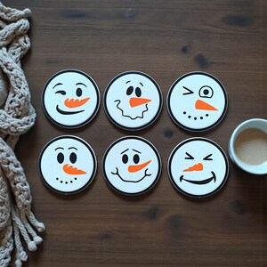 May include: Six round white coasters with snowman faces, each with a different expression. The snowmen have black eyes, orange carrot noses, and black smiles. The coasters have a black border and are arranged on a brown wooden surface.