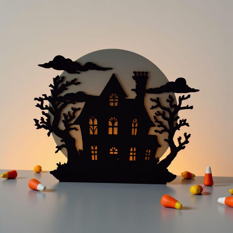 May include: A black silhouette of a haunted house with lit windows, trees, and clouds, set against a full moon. Candy corn is scattered in the foreground, creating a spooky Halloween scene.