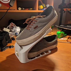 May include: A pair of gray canvas sneakers with brown laces and a white sole, resting on a white shoe rack. The shoes are worn and show signs of use. The rack is designed to hold two pairs of shoes.