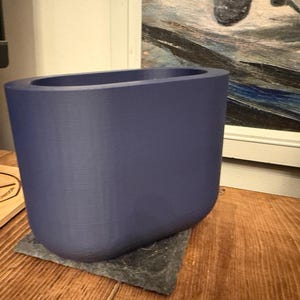 May include: A solid, indigo-colored planter with a rounded rectangular shape. The planter sits on a dark gray square base. The background includes a wooden surface and a framed painting.