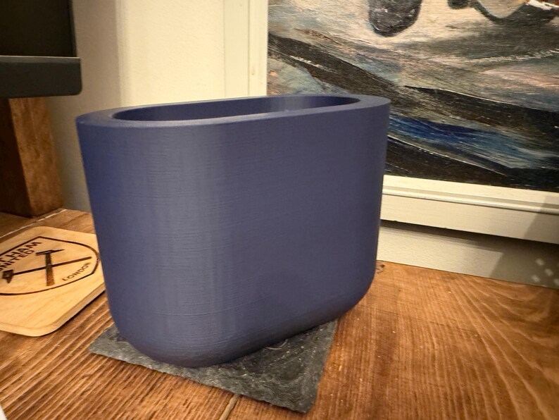 May include: A solid, indigo-colored planter with a rounded rectangular shape. The planter sits on a dark gray square base. The background includes a wooden surface and a framed painting.