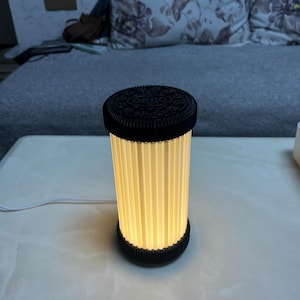 Pode incluir: Uma l&acirc;mpada cil&iacute;ndrica com um design de biscoito Oreo preto na parte superior e inferior. O corpo da l&acirc;mpada &eacute; uma s&eacute;rie de pain&eacute;is verticais plissados, esbranqui&ccedil;ados, que emitem um brilho quente. Um cabo de alimenta&ccedil;&atilde;o branco se estende da base.
