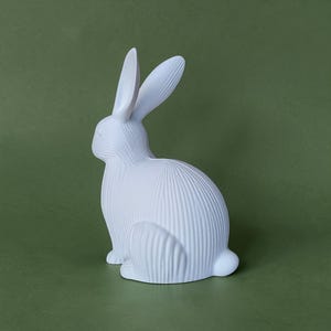 May include: A white ceramic rabbit figurine with textured vertical lines. The rabbit is in a seated position with its ears upright. The figurine is set against a solid green background. This decorative item is suitable for home decor.