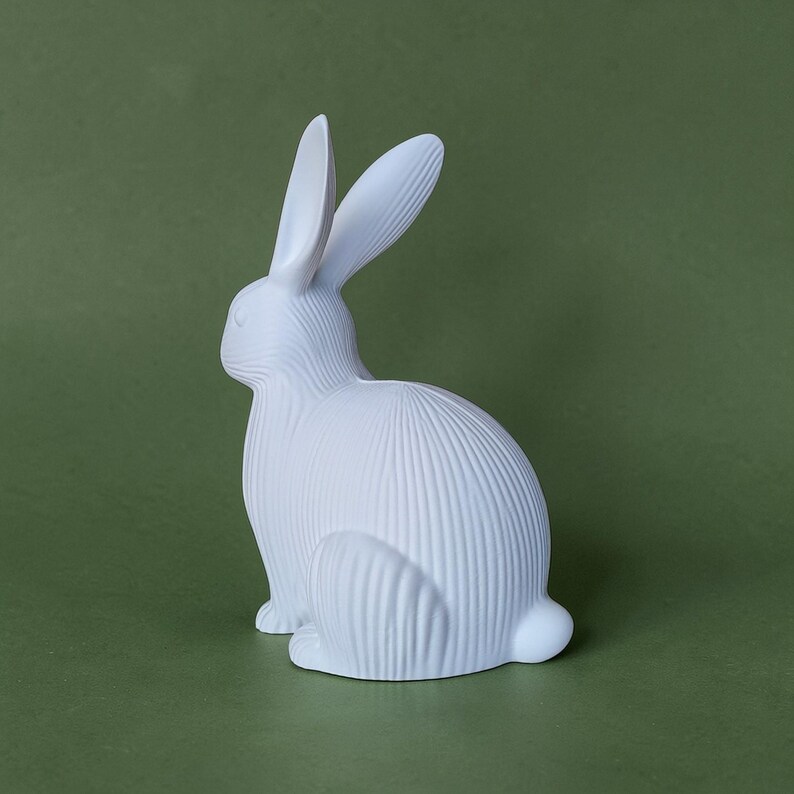 May include: A white ceramic rabbit figurine with textured vertical lines. The rabbit is in a seated position with its ears upright. The figurine is set against a solid green background. This decorative item is suitable for home decor.