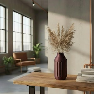 May include: A burgundy vase with vertical grooves holds dried ornamental grass. The vase sits on a wooden table with a stack of books to the right. The room has large windows and a leather armchair.