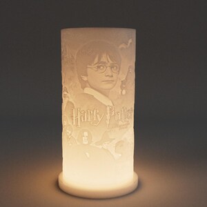May include: A cylindrical, off-white lamp featuring a portrait of Harry Potter, with the text "Harry Potter" and other imagery. The lamp is illuminated from within, casting a warm glow. The base is a simple, round design.