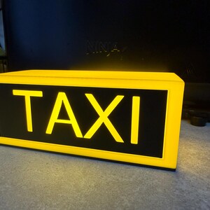 May include: Illuminated yellow taxi sign with the word "TAXI" in bold yellow letters on a black background. The sign has a rectangular shape and is lit from within, creating a bright glow.