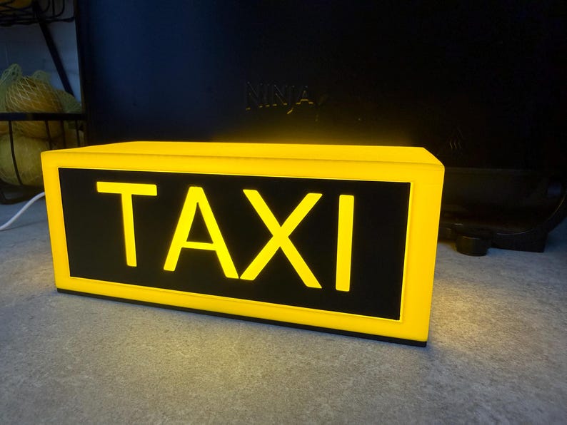 May include: Illuminated yellow taxi sign with the word "TAXI" in bold yellow letters on a black background. The sign has a rectangular shape and is lit from within, creating a bright glow.