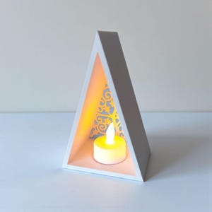May include: A white, triangular candle holder with a decorative cutout design on the back panel. Inside, a small, lit, yellow candle sits on a light pink base, creating a warm glow. The candle holder is a modern home decor item.
