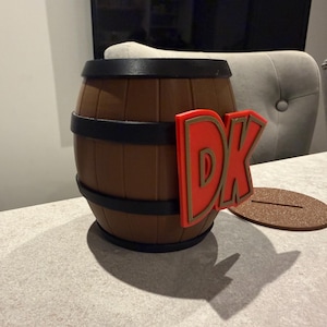 May include: A brown barrel with black bands and a red and gold "DK" logo. The barrel sits on a light-colored surface with a brown, oval-shaped base. The barrel is a decorative item.