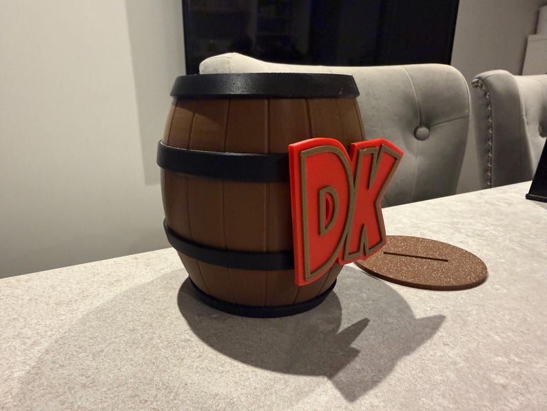 May include: A brown barrel with black bands and a red and gold "DK" logo. The barrel sits on a light-colored surface with a brown, oval-shaped base. The barrel is a decorative item.