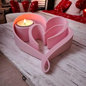 May include: A pink heart-shaped candle holder with a lit candle. The holder has a layered design with a circular space for the candle and a decorative heart shape. The candle holder is on a light-colored wooden surface.