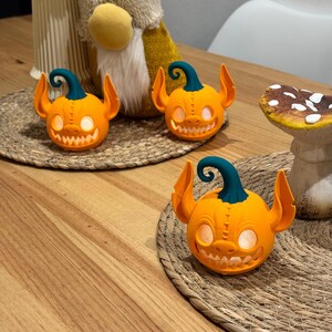 May include: Three orange pumpkin-shaped Halloween decorations with teal stems and ear-like protrusions. Each pumpkin has a carved, toothy grin and glowing eyes. They sit on a woven placemat, suggesting a festive autumn theme.