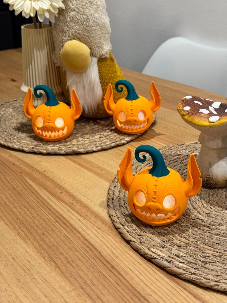 May include: Three orange pumpkin-shaped Halloween decorations with teal stems and ear-like protrusions. Each pumpkin has a carved, toothy grin and glowing eyes. They sit on a woven placemat, suggesting a festive autumn theme.