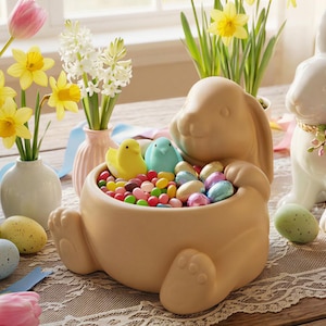 May include: A beige ceramic bunny-shaped container filled with colourful Easter candies and two small bird figurines. The container is surrounded by decorative Easter eggs, flowers, and a white ceramic bunny with a floral collar.