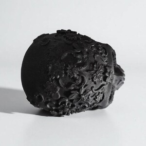 May include: Black decorative skull with floral patterns. The skull is made of a matte black material and features detailed, raised floral designs across its surface. The skull is positioned on a white background.
